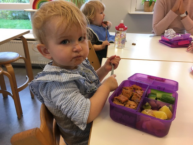 En lille pige sidder ved et bord med hagesmæk på og sin madkasse foran sig. Foto: Frederikssund Kommune.