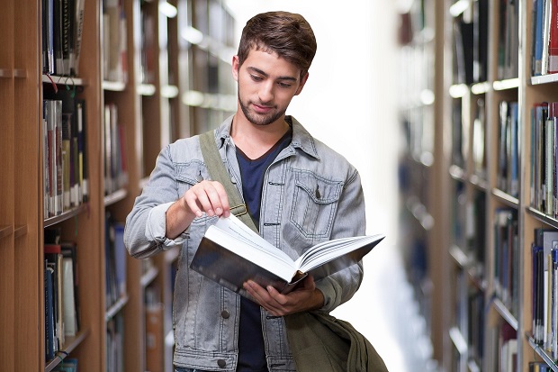 Ung mand læser i bog mens han står  mellem reoler med bøger. Foto: Pixabay