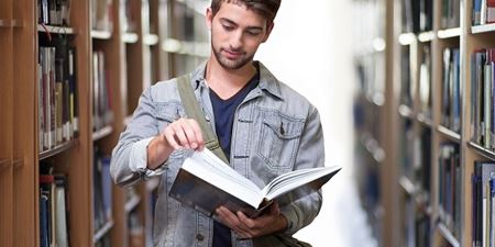 Ung mand læser i bog mens han står  mellem reoler med bøger. Foto: Pixabay