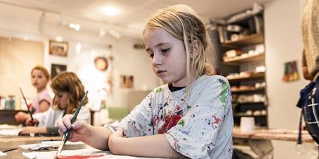 Pige sidder og maler og er iført en hvid t-shirt med en masse malerpletter på. Foto: Frederikssund Kommune, Kenneth Jensen.