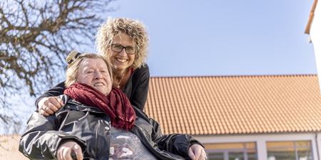 Ældre kvinde i rullestol skubbes frem af en anden yngre kvinde. Begge smiler og solen skinner fra en blå himmel. Foto: Frederikssund Kommune, Kenneth Jensen.