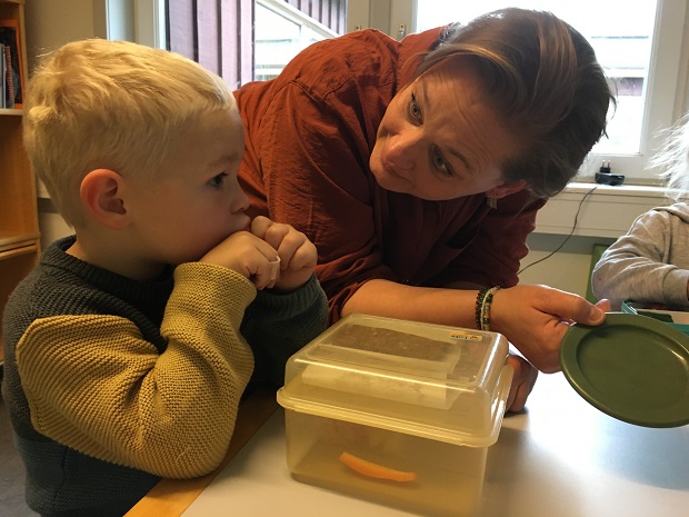 En dreng sidder ved et bord med sin madpakke og en voksen, Sarah Kofoed Hvid, står bøjet ned ved siden af ham med en legetallerken i hånden. Foto: Frederikssund Kommune.